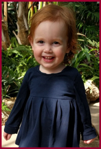 A young girl with red hair smiling in front of a plant. Her name is Alice.