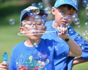 A little boy in a blue shirt and hat blows bubbles as another boy watches from behind