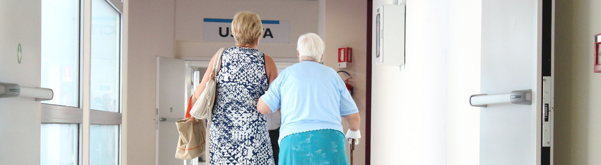An older woman walks her elderly mother through the bright hallway of a medical facility