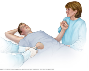 An illustration showing a patient receiving a liver biopsy. The person lays on a table, holding hands with a visitor while a doctor inserts a needle into the patient's right side. Image used with permission by Mayo Clinic