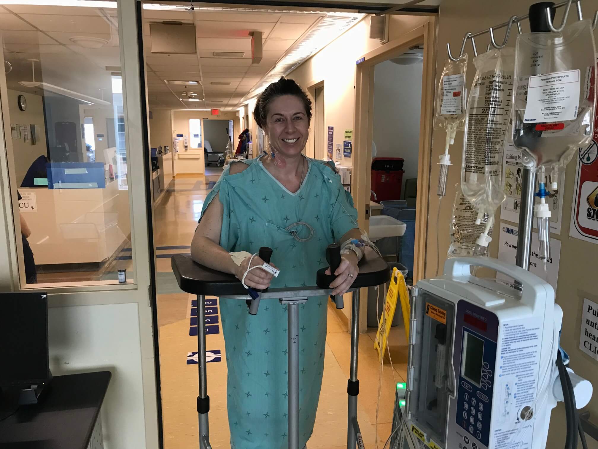 Heather leans on a walker smiling. She is wearing a hospital gown and is connected to a machine with IV bags.