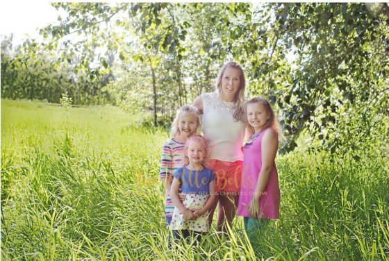Marla and her three young daughters, elementary school age, stand in a field surrounded by trees and wild grass.