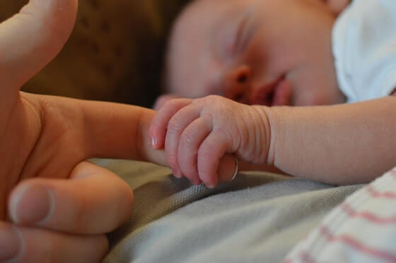 A baby holds an adults index finger while it sleeps.