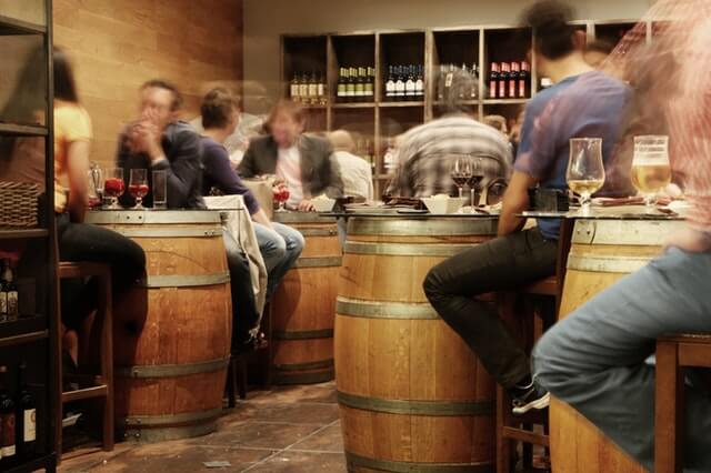 A blurry image of a bar depicts 2 to 3 guests dining on wooden barrels that have been converted to tables. Each table has some sort of alcoholic beverage on it, (wine and beer). The blur is most likely used to show that the viewer is intoxicated.