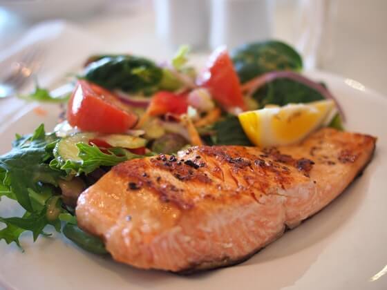 A piece of grilled salmon sits on a bed of salad. Visible are also tomato, sliced brussel sprouts, onions and a lemon wedge. 