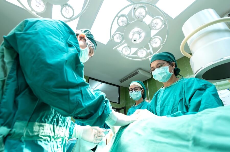 A team of surgeons operate on a patient who cannot be seen. Large lamps look down above them. 