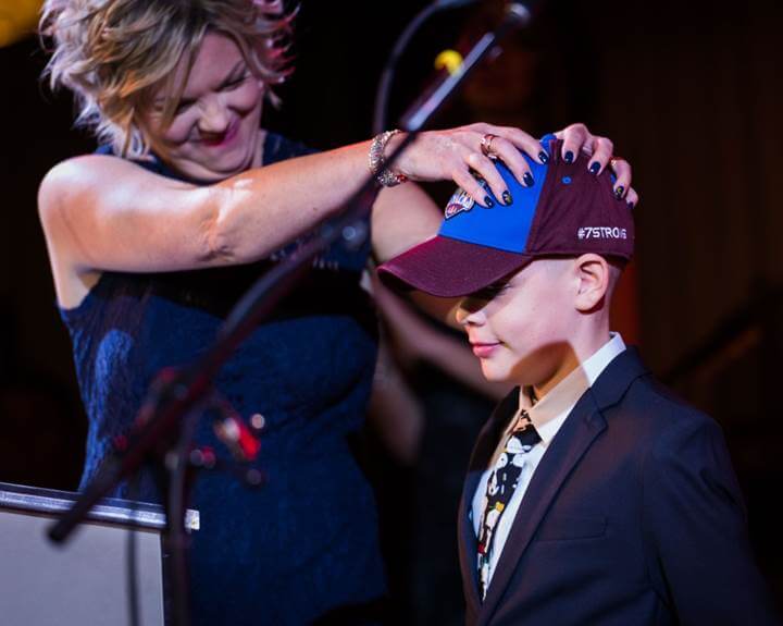 Brock chessell stands beside his mom in a suit and baseball cap. Julie is softly pressing her hands on Brock's head while smiling and holding back tears.