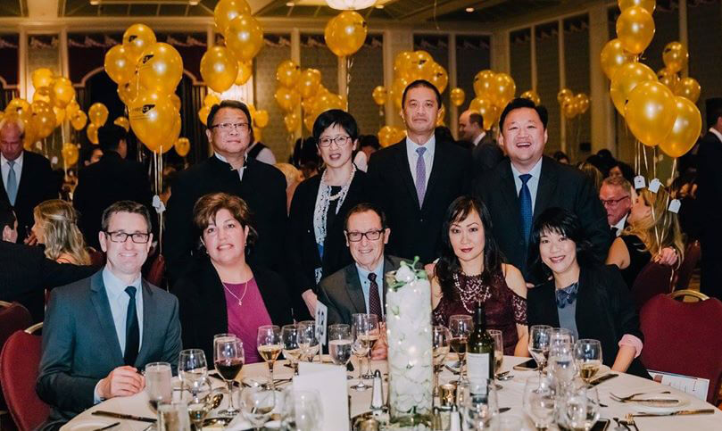 Attendees of the Gala smile for the camera, some seated at a table and some standing behind there. In the background there are golden balloons with the Canadian Liver Foundation logo printed on them