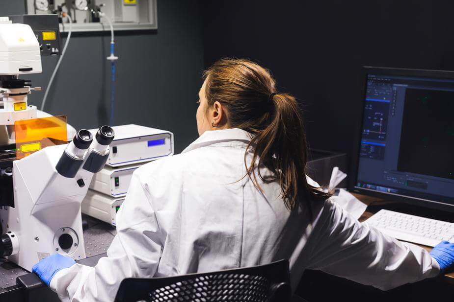 A female researcher sits in a lab with her back to the camera. She is wearing a lab coat with blue gloves. Her left hand is positioned on a microscope while her right hand is on a key board. More equipment is stationed around the room.