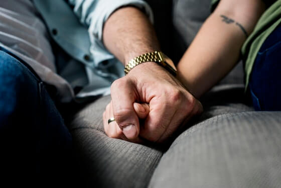 A man and woman hold hands while sitting on a couch, only their arms are visible. The man has a gold watch on. The woman has a tattoo on her forearm and appears to be wearing a wedding ring. One of these partners are presumably the caregiver for their loved one.