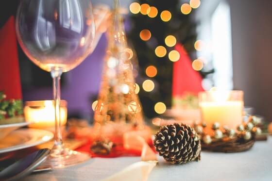 A table is set for the holiday season. In the background, a christmas tree with bright lights are out of focus. On the table, there two lit candles, a red table cloth, some gold beeds, a goald painted pine cone and a wine glass with a dinner plate visible.