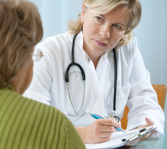 A doctor listens attentively while taking notes of what her patient is telling her. She is wearing a stethoscope and writing with pen on a clipboard. The patient is a middle-aged woman her glasses, and her
