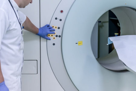 A man stands to the side of a large machine with an opening at the center. The man is wearing a labcoat and blue latex gloves. His left hand is resting on a panel of buttons that control the machine.