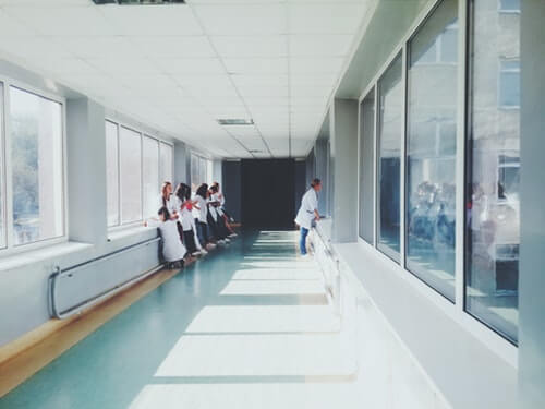 A hallway in the hospital with windows to outside facing on the left, and windows into rooms facing on the right. There are 5 woman leading up against the walls, presumably doctors in training while another older-looking female doctor peers into the room windows.
