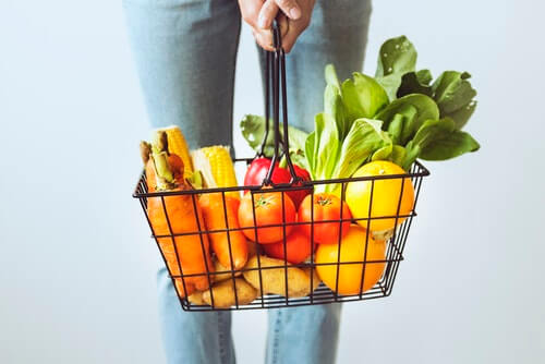 A woman holds a shopping basket with colourful fruits and veggies like carrots, corn, red peppers, lettuce, tomatoes, and oranges.