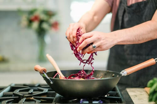 A man is seperating shredded red cabbage into a frying pan with a wooden handle and a wood spoon sitting in the pan. The heat is on and some other ingredients can be slightly seen. In the background, a vase full of red flowers can be seen out of focus, which may paint the picture of a Valentine's day dinner