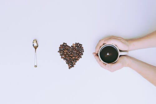 From left to right, a teaspon, coffee beans arranged in a heart, and a person holding a coffee cup with their hands wrapped around them are shown. The background is blank and grey. 