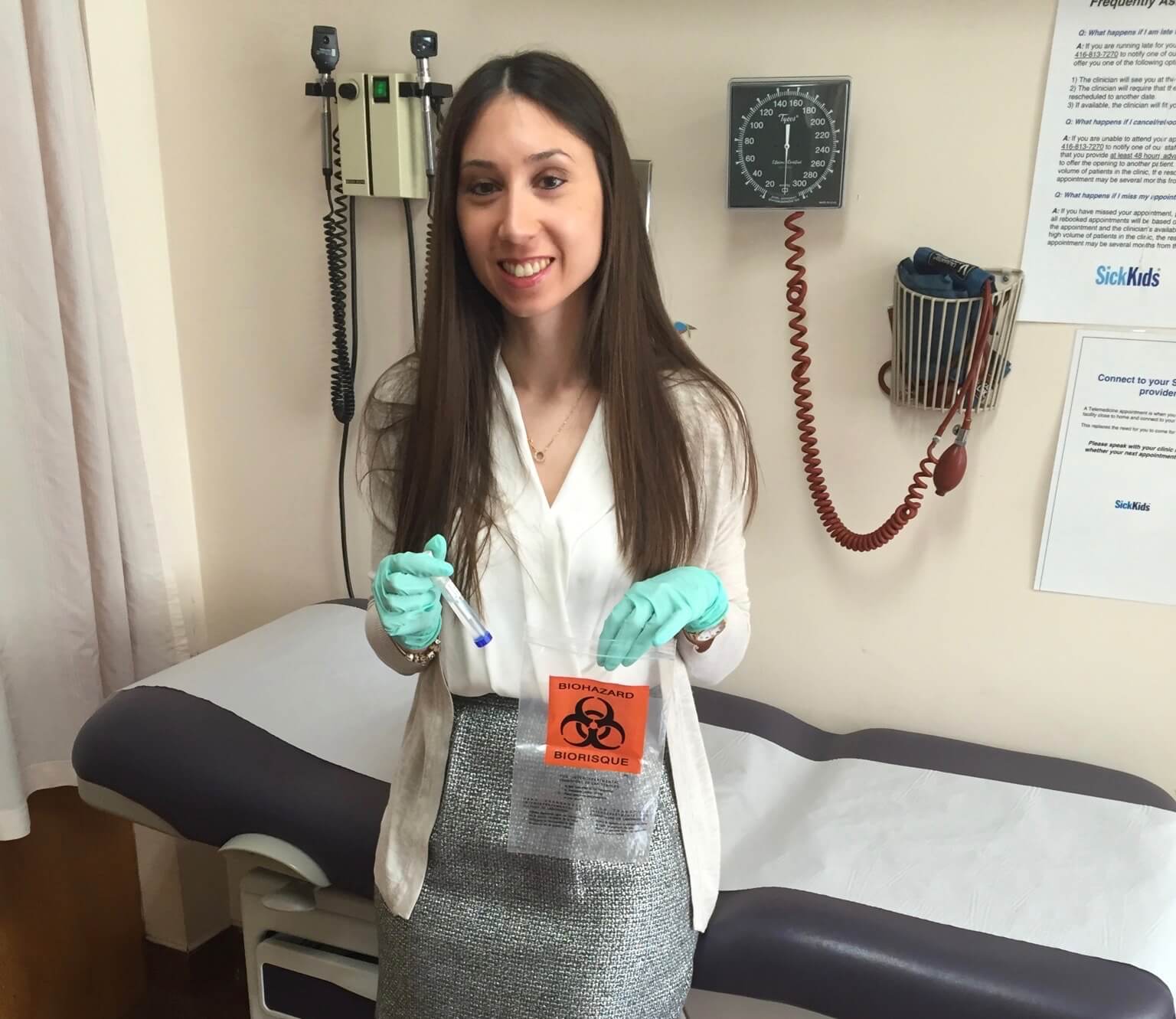 Amanda stands in a hospital room with gloves on and holding a test tube and plastic bag. It can be concluded from the photo that she is holding a sample from her PSC IBD study that will be carefully examined in a lab at a later date.