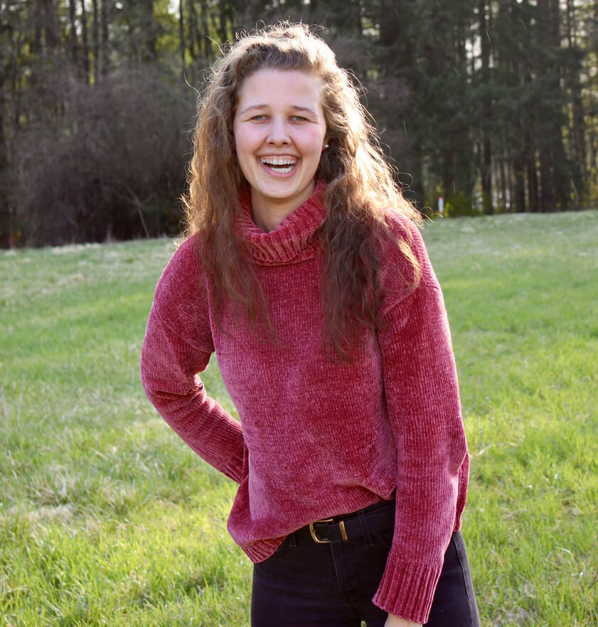 Natalie poses in a grassy field with a forrest in the backdrop. She is wearing a dark red turtle neck styled sweater and black jeans. 