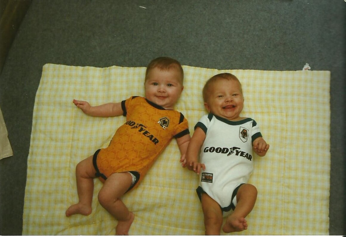 Natalie and her sister as infants lay on their backs on a yellow checkered blanket. They are both in Good Year branded onesie's, one orange and one white. 