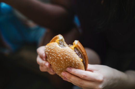 A male, not pictured, is holding a cheeseburger and sitting on a couch. The background is blurred so that the cheesburger and hands are in focus