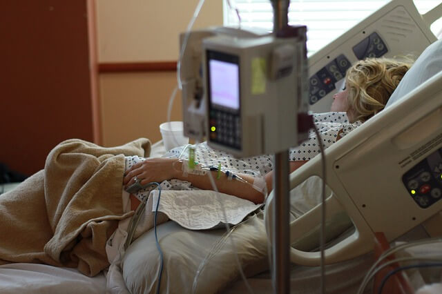 A woman lies in a dark hospital room with her bed slightly elevated. Her left arm has an IV in it and a number of machines surround her.