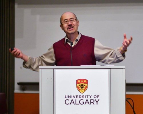 A doctor speaks at the Canadian Liver Foundation's LIVERight Forums, He is standing in front of a podium in mid sentence with his hands positioned outwards. He is wearing a dark red vest with a beige dress shirt underneath. In his right hand, he holds a remote for a PowerPoint presentation (not pictured). The podium has the logo of the University of Calgary branded on it. 