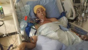 Heather lies in a bed connected to all sorts of tubes and medical apparatuses one would imagine are keeping her alive. She has an oxygen tube and an IV that is visible. A yellow wash cloth lies over her forehead.
