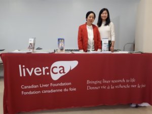 Andrea stands behind a table that has a Canadian Liver Foundation branded table cloth. She is posing with another volunteer, who along with Andrea, is surrounded by informational brochures on liver disease and liver health. 
