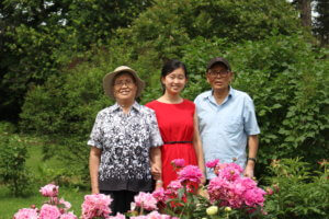 A teenage Andrea stands in the middle of her grandparents smiling in what looks to be a garden. Light and deep pink flowers are pictured at the bottom of the frame, with dark green trees and bushes surrounding the three of them. 