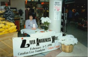 A Canadian Liver Foundation Volunteer from Calgary sits at a display booth in a shopping mall. The table of the booth has a banner that state "liver awareness week", and pamphlets can be seen on the table.