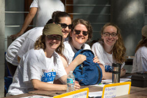 A group of young ladies are seated at a registration table at a Stroll for liver devent. They are volunteering to help register people for the event and get them the gear and information they may need. 