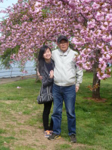 Andrea, now a young adult, stands in front of a large cherry blossom tree with her grandfather. He has his arm around her, and is wearing a light grey windbreaker with a dark hat. In the background, a body of water can be seen. 