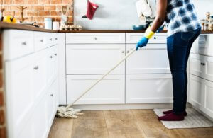 A person stands on a kitchen map and extends a mop over towards kitchen cabinets. He or she is wearing blue rubber gloves.