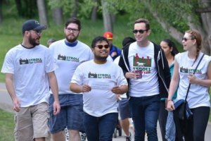 A group of friends in their early adult years walk down a park path talking and smiling. Pictured, are their Stroll for Liver t-shirts. Some of them wear sunglasses.