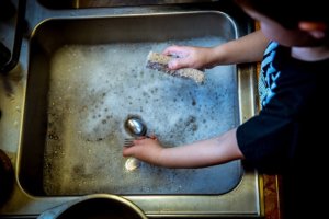 A child stands over a sink that is filled with soapy waters. He is holding a sponge in one hand and some utensils in the other, looking ready to scrub and wash these dishes.