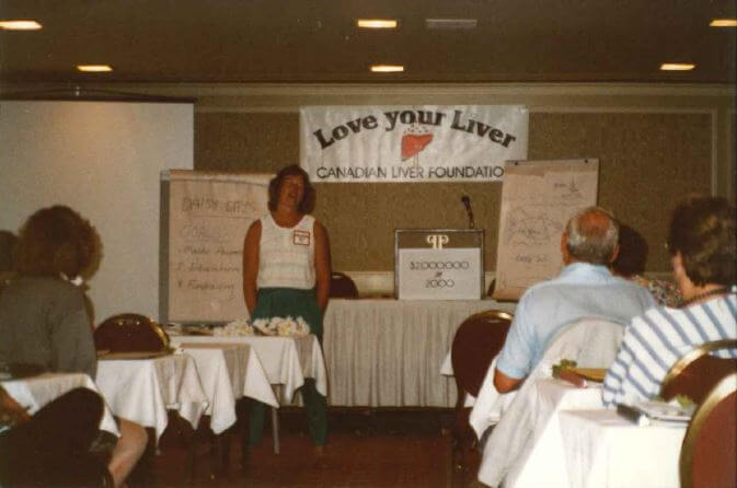 Annette presents to a room full of volunteers at an educational event in Calgary. Behind her, a banner reads "love your liver!" and signage can be seen. 