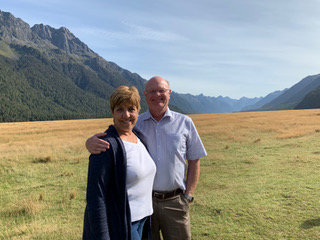 A man and woman pose for a photo in a green filled infront of mountains